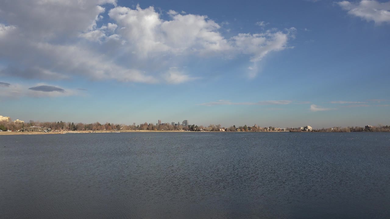 Wide Westminster, Colorado water view with rippled foreground, low shoreline, distant buildings, large white clouds, and no visible people.