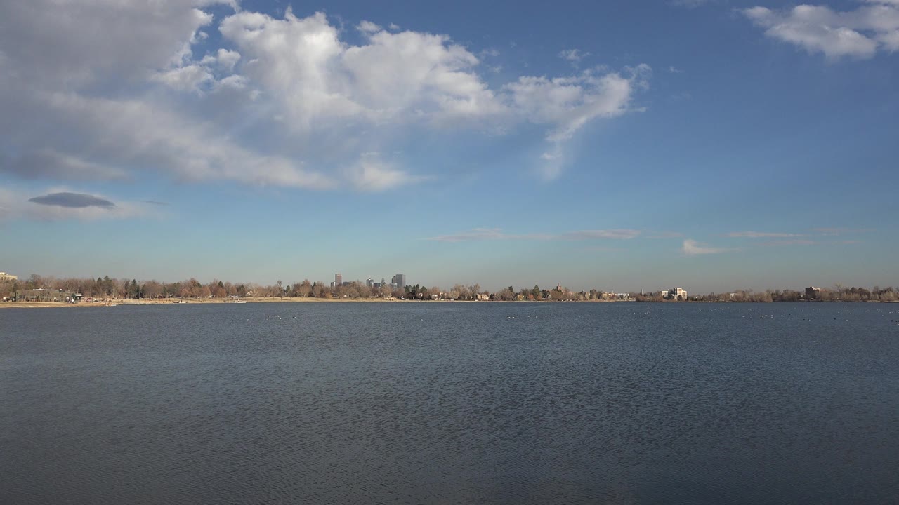 Opening frame of a Westminster, Colorado water landscape with rippled blue water, a low tree-lined shoreline, large white clouds, and no visible people.