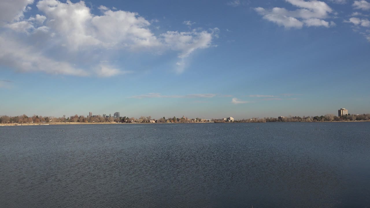 Opening frame of Westminster, Colorado water with ripples in the foreground, a low distant shoreline, scattered buildings, large clouds overhead, and no visible people.
