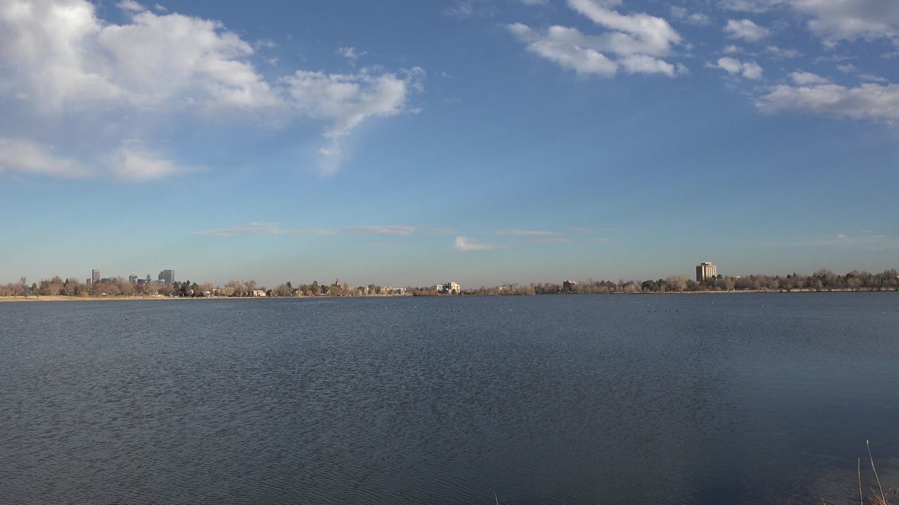 Opening frame of a wide Westminster, Colorado water view with rippled surface, distant shoreline trees and buildings, scattered white clouds, and no visible people.