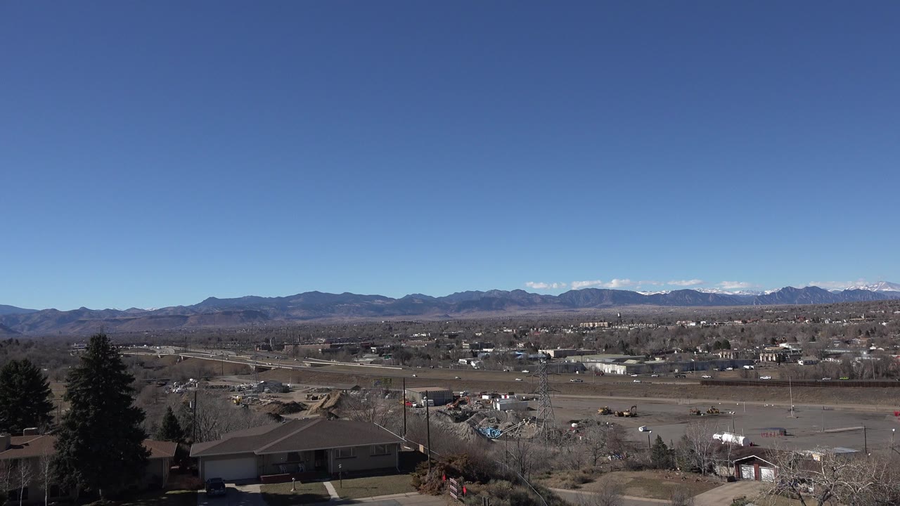 Wide daylight view over Westminster, Colorado with foreground rooftops and trees, roads and scattered cars in the midground, distant mountains, and no visible people.