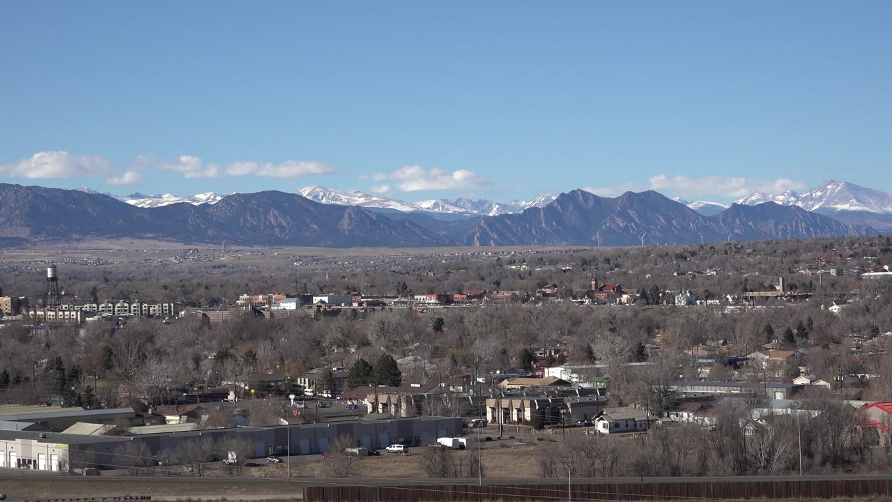 Wide view of Westminster, Colorado with low buildings, leafless trees, distant snow-touched mountains, clear blue sky, and no visible people.