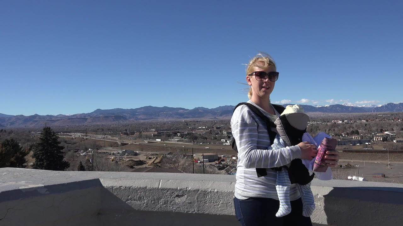 The woman remains near the left side of the frame holding the baby and a purple cup, with the low wall, open land, roads, and mountain horizon visible in Westminster, Colorado.