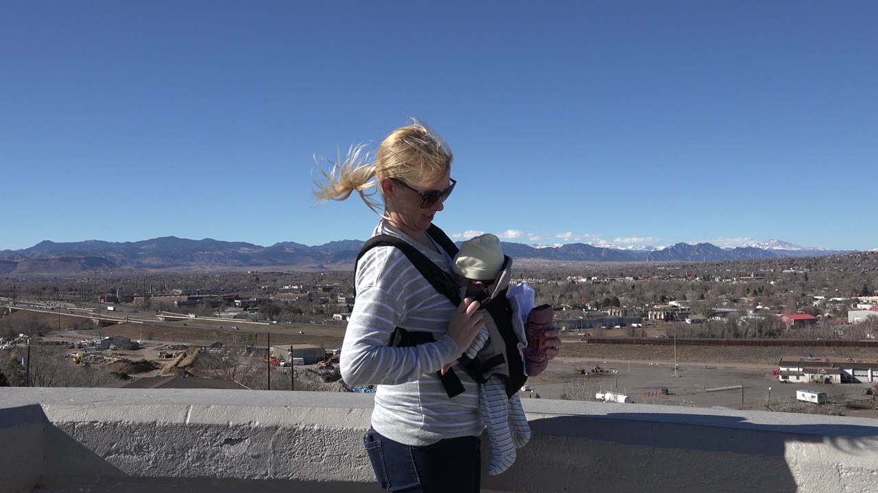 Mother in sunglasses looks down at baby in a front carrier beside a concrete overlook.