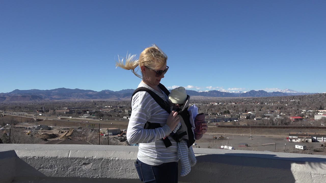 At 0:01 in Westminster, Colorado, the mother remains turned toward the infant carrier, with her hair blown sideways and the broad suburb-to-mountain view behind her.