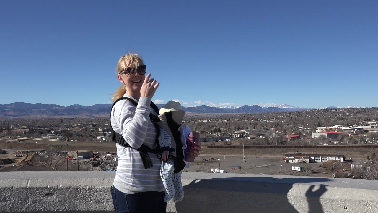 In Westminster, Colorado, a blonde woman wearing sunglasses carries an infant in a front carrier beside a concrete ledge with neighborhoods and mountains behind her.