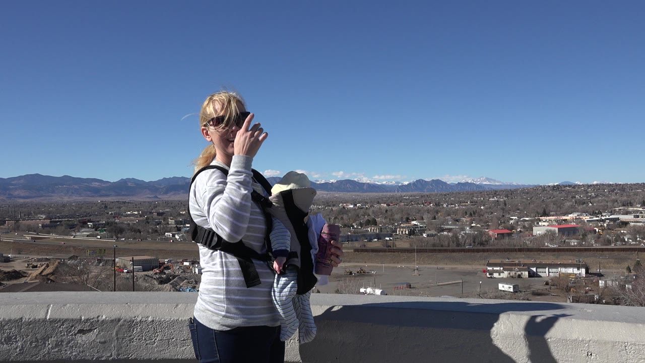 The mother raises one hand near her face while the infant rests in the front carrier, with Westminster rooftops, open land, and mountains stretching across the background.
