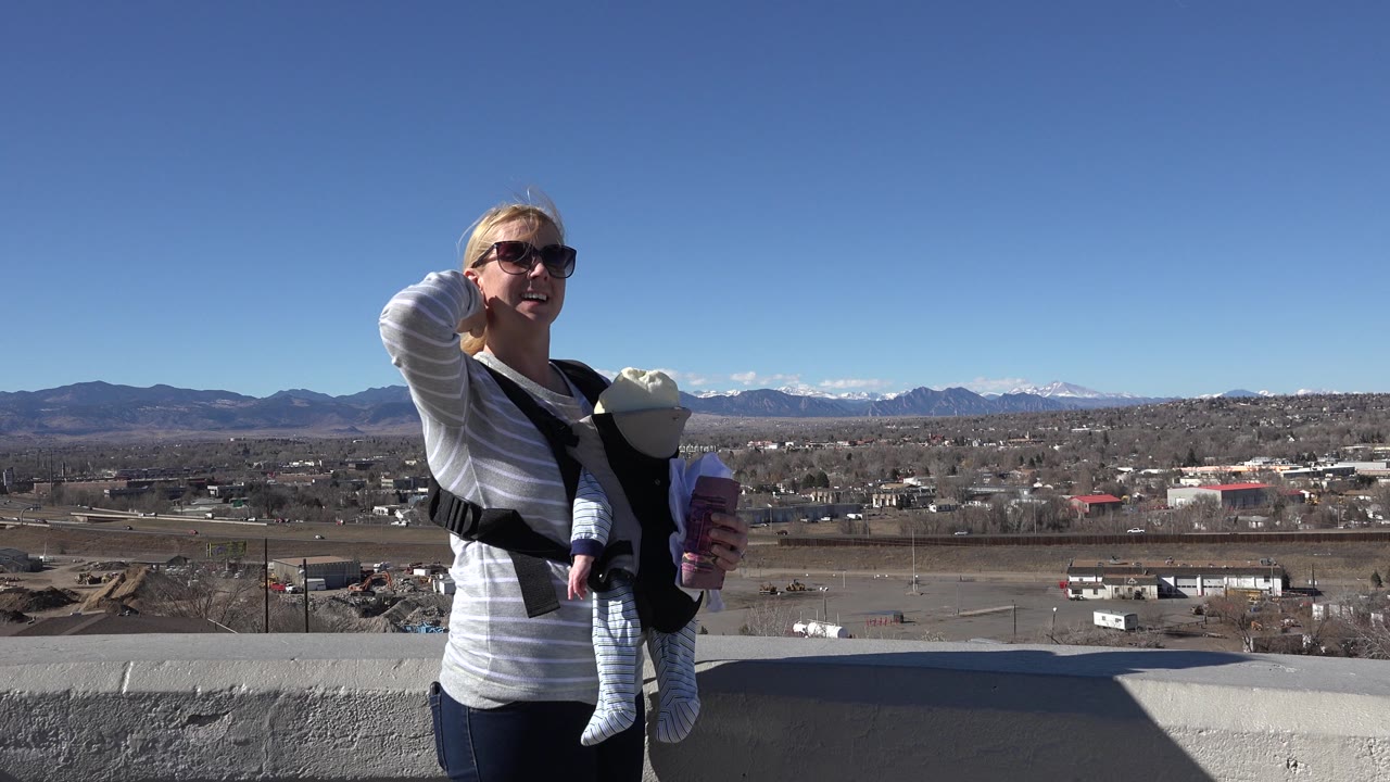 Woman carrying a baby touches her hair in front of a wide Colorado landscape.
