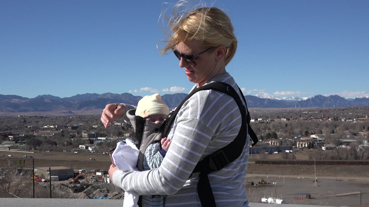 Blonde woman in sunglasses holding a baby in a front carrier outdoors.
