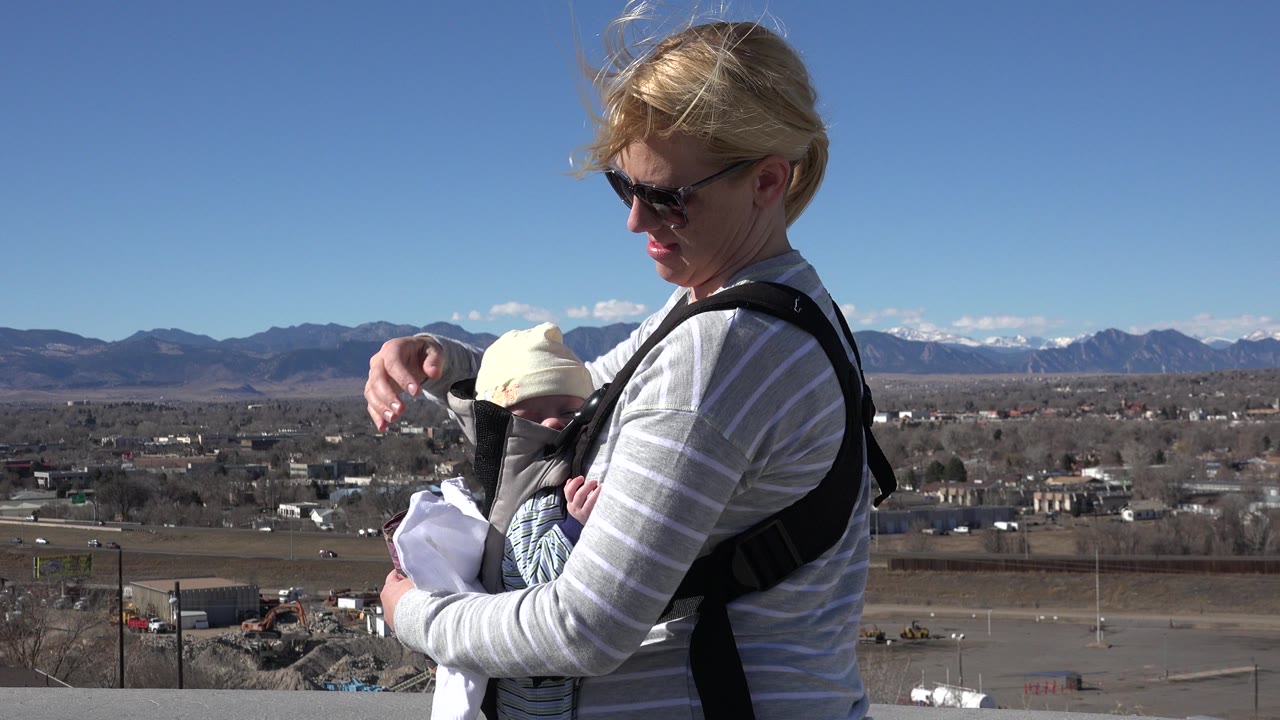 One second in, the woman keeps her gaze on the infant in the carrier, with wind moving her hair and the Westminster road and mountain view behind them.