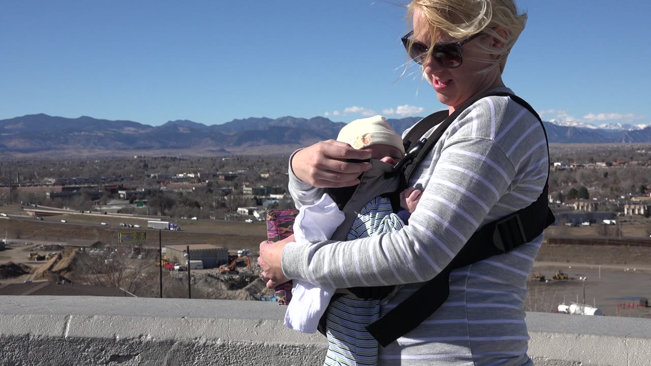At 0:01, the woman looks down toward the infant while standing near a concrete overlook in Westminster, Colorado, with highway lanes and a mountain ridge in the distance.