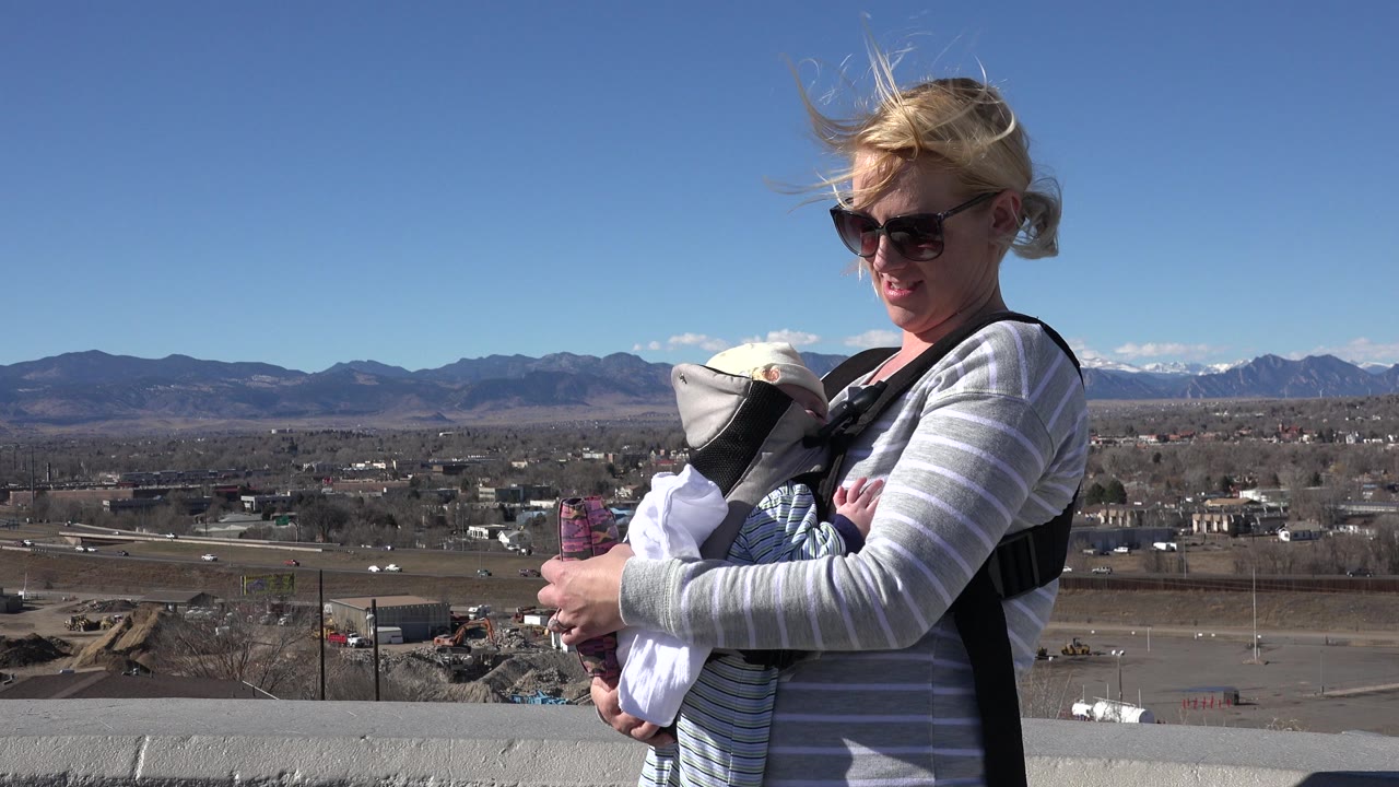 Blonde woman in sunglasses holds a baby in a front carrier at an overlook in Westminster, Colorado, with distant snow-capped mountains behind her.