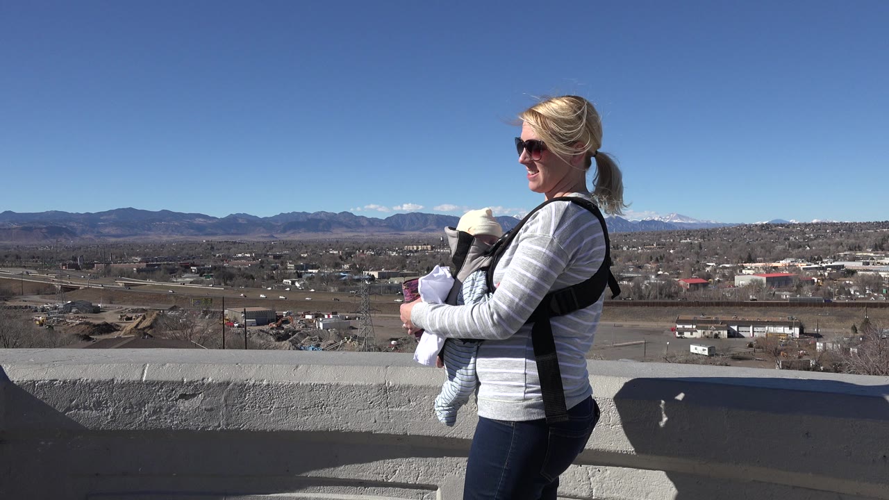 Woman in sunglasses carrying a baby beside a concrete overlook wall.