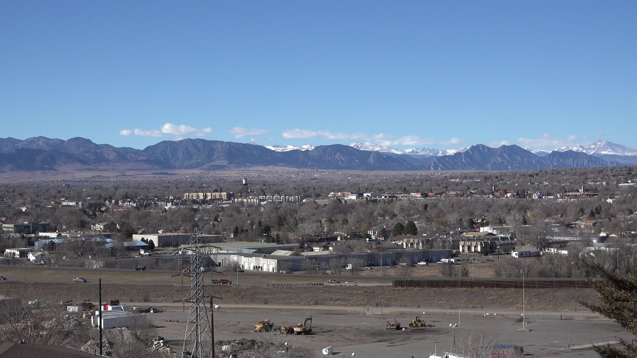 Wide view across Westminster, Colorado buildings, roads, utility towers, bare trees, distant mountains, and a clear blue sky with small white clouds, with no visible people.