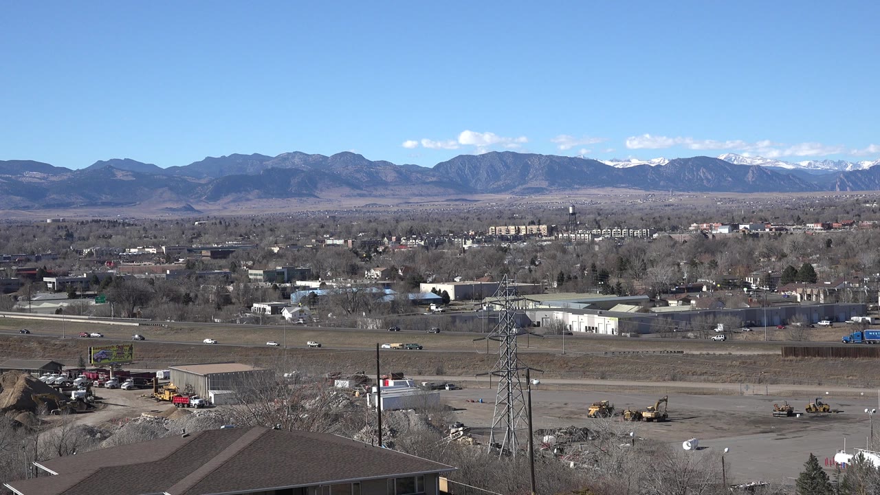 Wide first frame of Westminster, Colorado with a roofline in the foreground, light traffic on the road, a utility tower, low buildings, distant mountains, and no visible people.