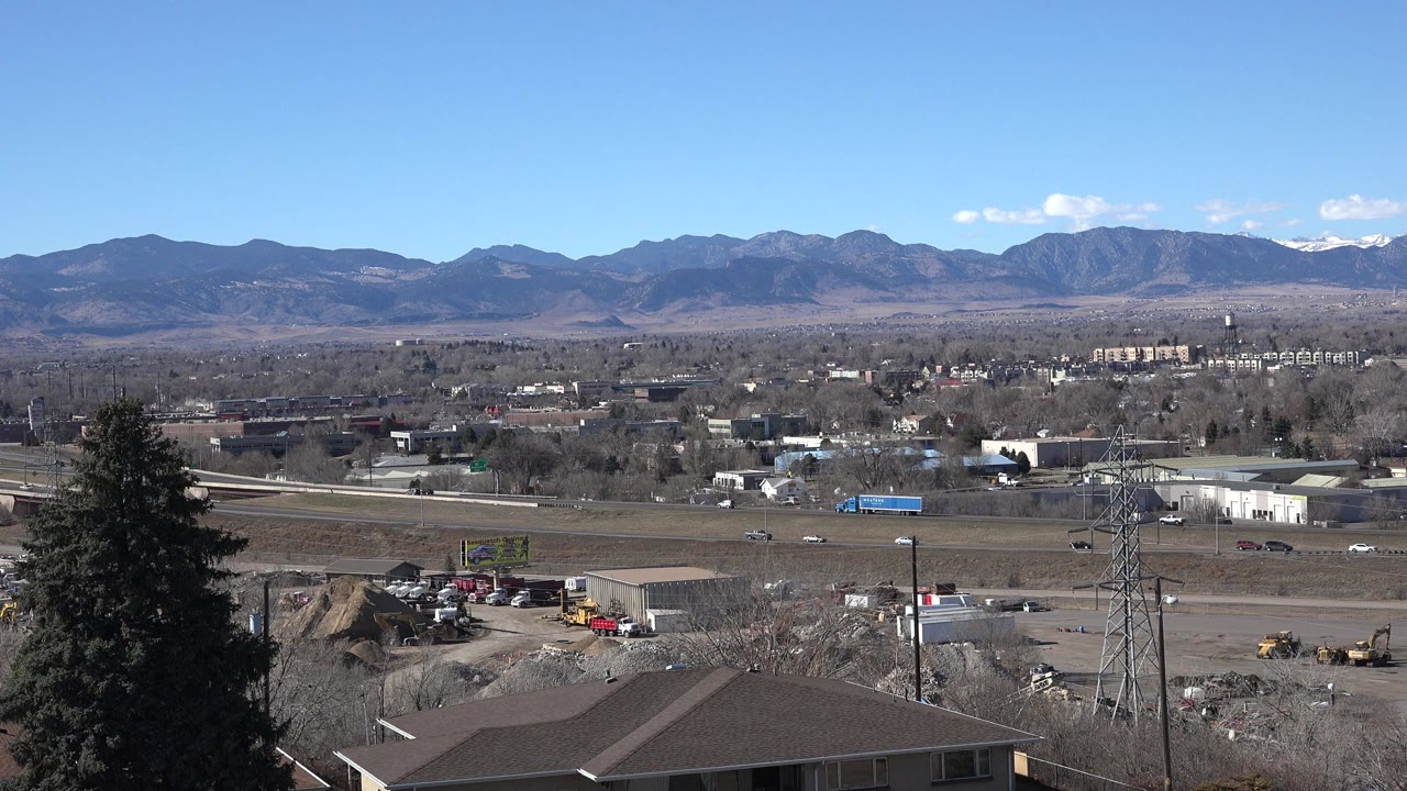 Opening frame of Westminster, Colorado from an elevated ground view, with a tall evergreen at left, rooftops below, light traffic on roads, distant mountains, and no visible people.