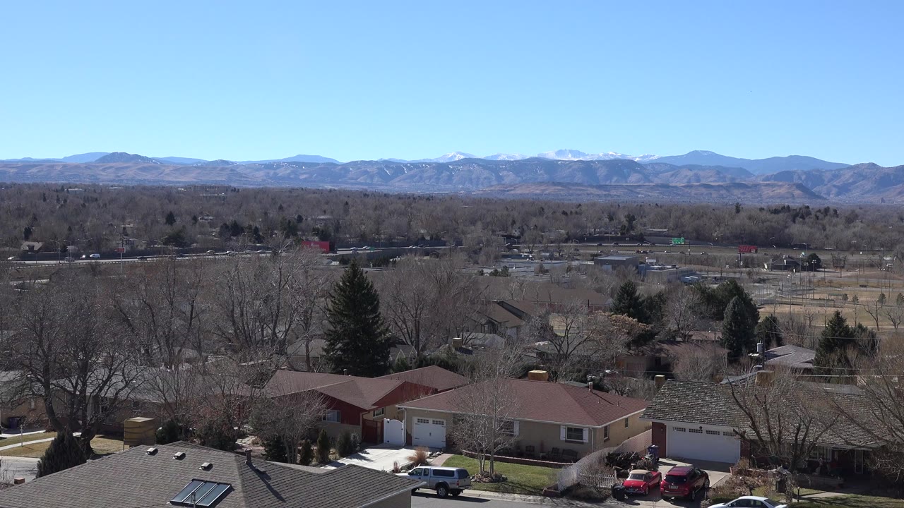 Opening view in Westminster, Colorado, showing private homes and rooftops below leafless trees, with roads, scattered cars, and distant mountains, no visible people.