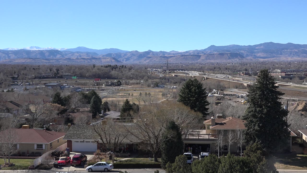 Wide suburban view with houses, trees, roads, and distant mountains under blue sky.