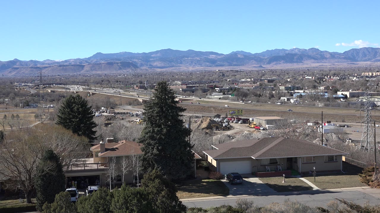 Westminster, Colorado frame showing foreground houses and bare trees, midground road lanes and work area, and mountains stretching across a clear blue sky.