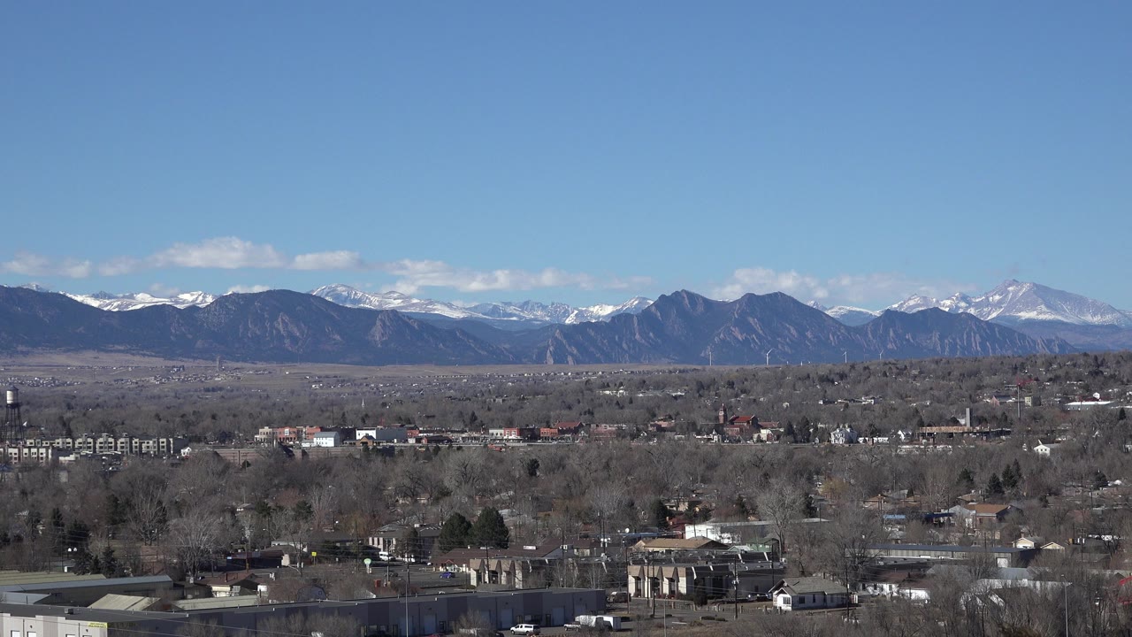 Opening view from Westminster, Colorado across low buildings, leafless trees, and utility structures toward snow-capped mountains under a clear blue sky, with no visible people.