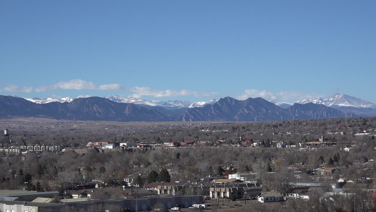 Westminster, Colorado appears from an elevated viewpoint with brown foothills, snow-tipped peaks, low commercial buildings, and bare winter trees, with no visible people.