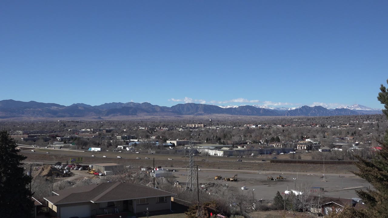 Opening 0:00 frame of Westminster, Colorado from an elevated hillside, showing rooftops, roads, trees, open land, distant mountains, and no visible people.