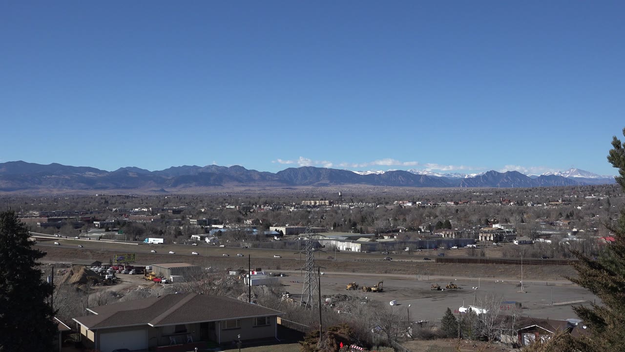 At 0:01, the Westminster, Colorado view holds a broad horizon with low buildings, open lots, rooftops, sparse trees, mountains, and no visible people.