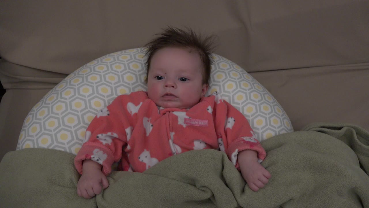 At 0:00, a brown-haired baby in Westminster, Colorado lies centered on a green blanket with a patterned pillow behind the head and a beige couch in back.