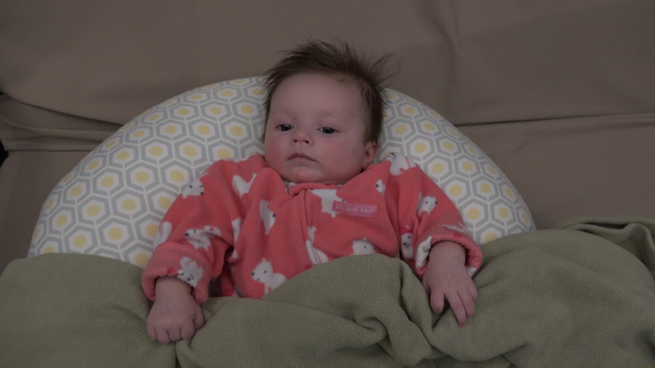 A baby in Westminster, Colorado lies against a gray-and-yellow patterned pillow with a green blanket pulled up, looking forward near the start of the clip.