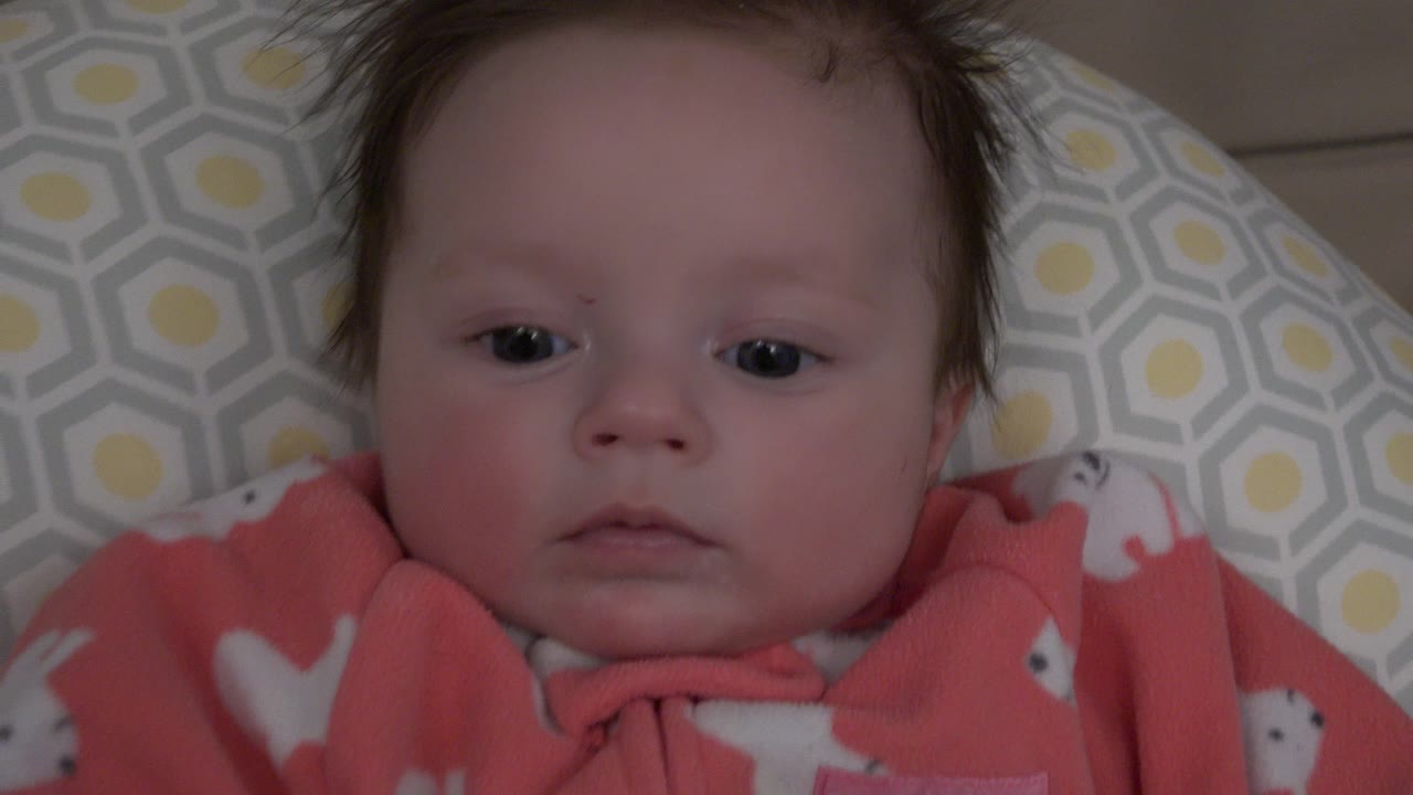 At 0:01, the infant remains centered on the patterned bedding, eyes open and mouth relaxed, with the coral outfit visible at the bottom of the frame.