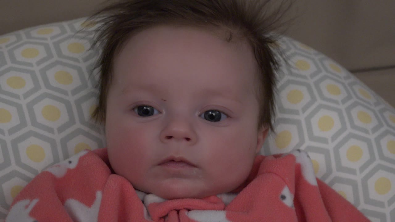 At 0:00, a baby in pink fleece looks straight toward the camera while lying on a gray, white, and yellow patterned cushion in Westminster, Colorado.