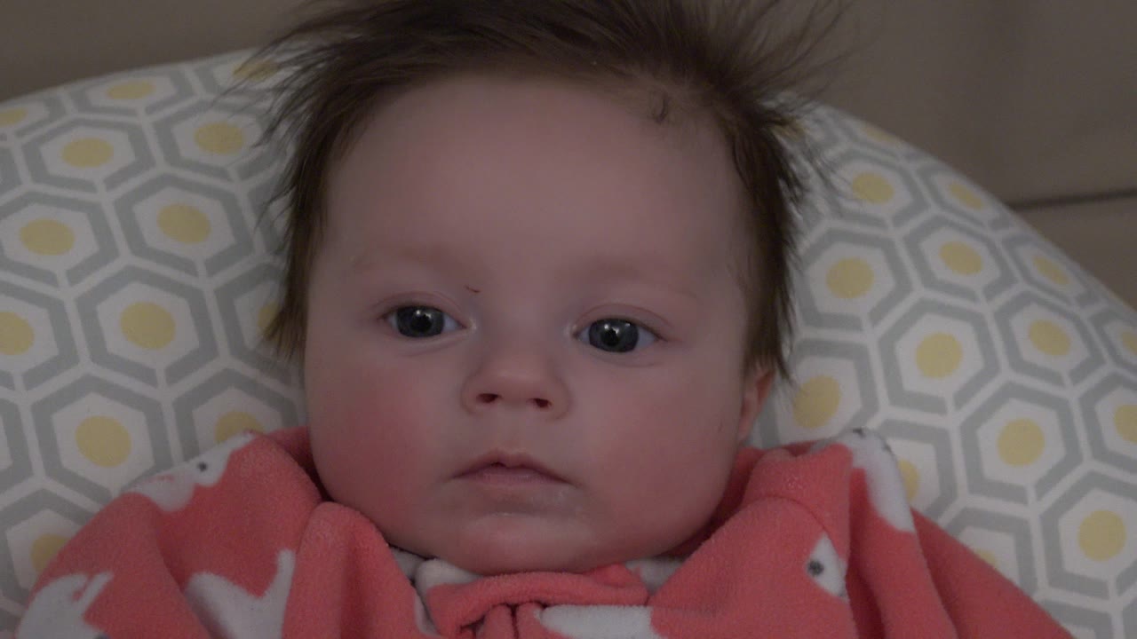 At 0:01, the baby remains centered in a tight close-up, eyes open toward the camera, with the patterned cushion filling the background in Westminster, Colorado.