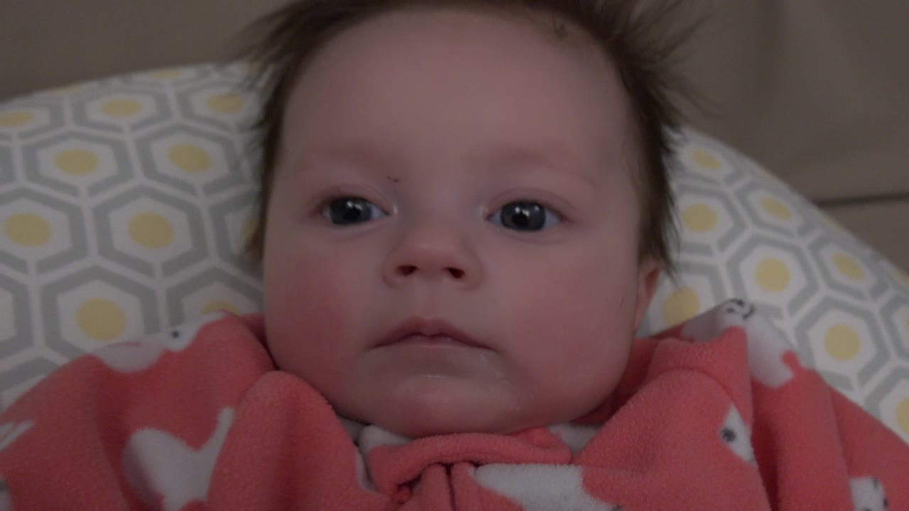 The baby remains centered in the Westminster, Colorado home frame, with dark eyes open, cheeks softly lit, and the coral clothing visible at the bottom edge.