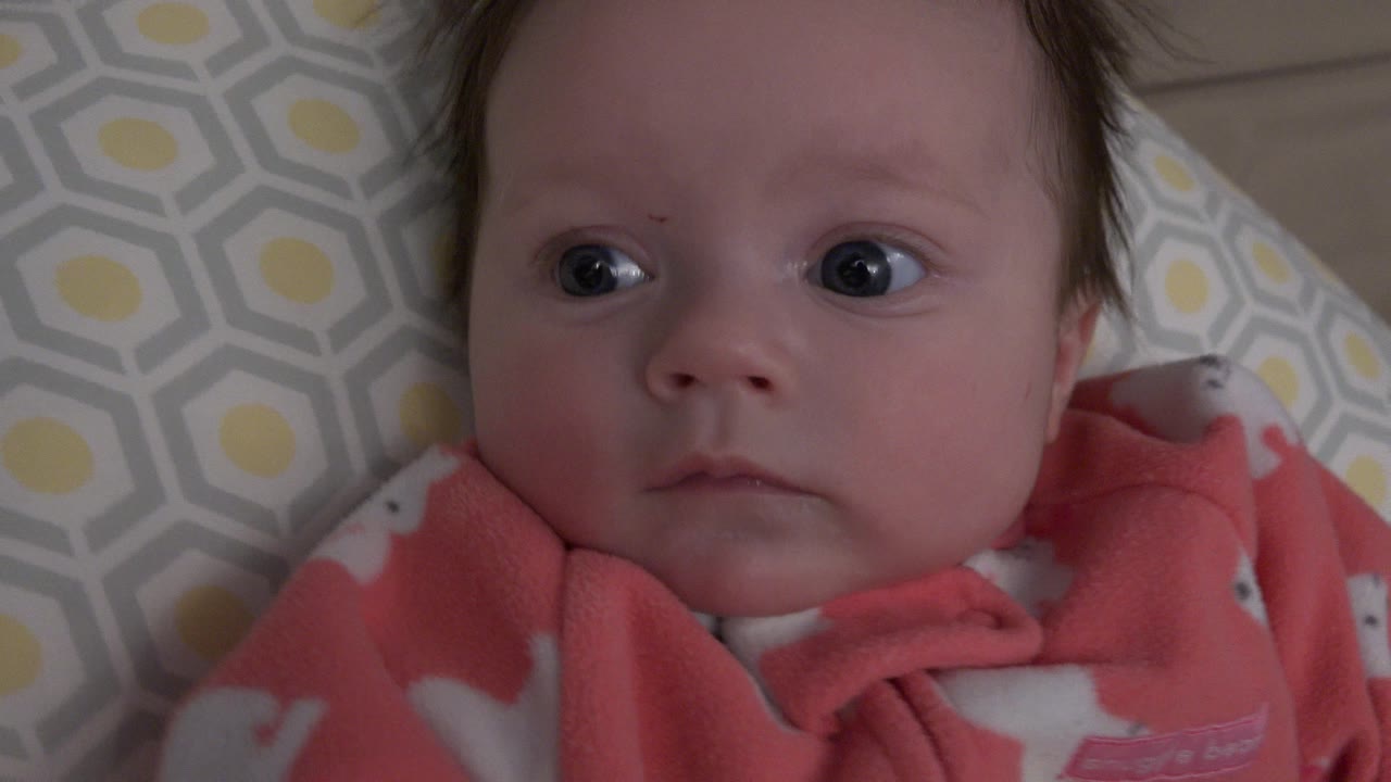 At 0:00, a baby in a pink fleece outfit lies on a pale blue and yellow patterned cushion in Westminster, Colorado, looking slightly off camera.