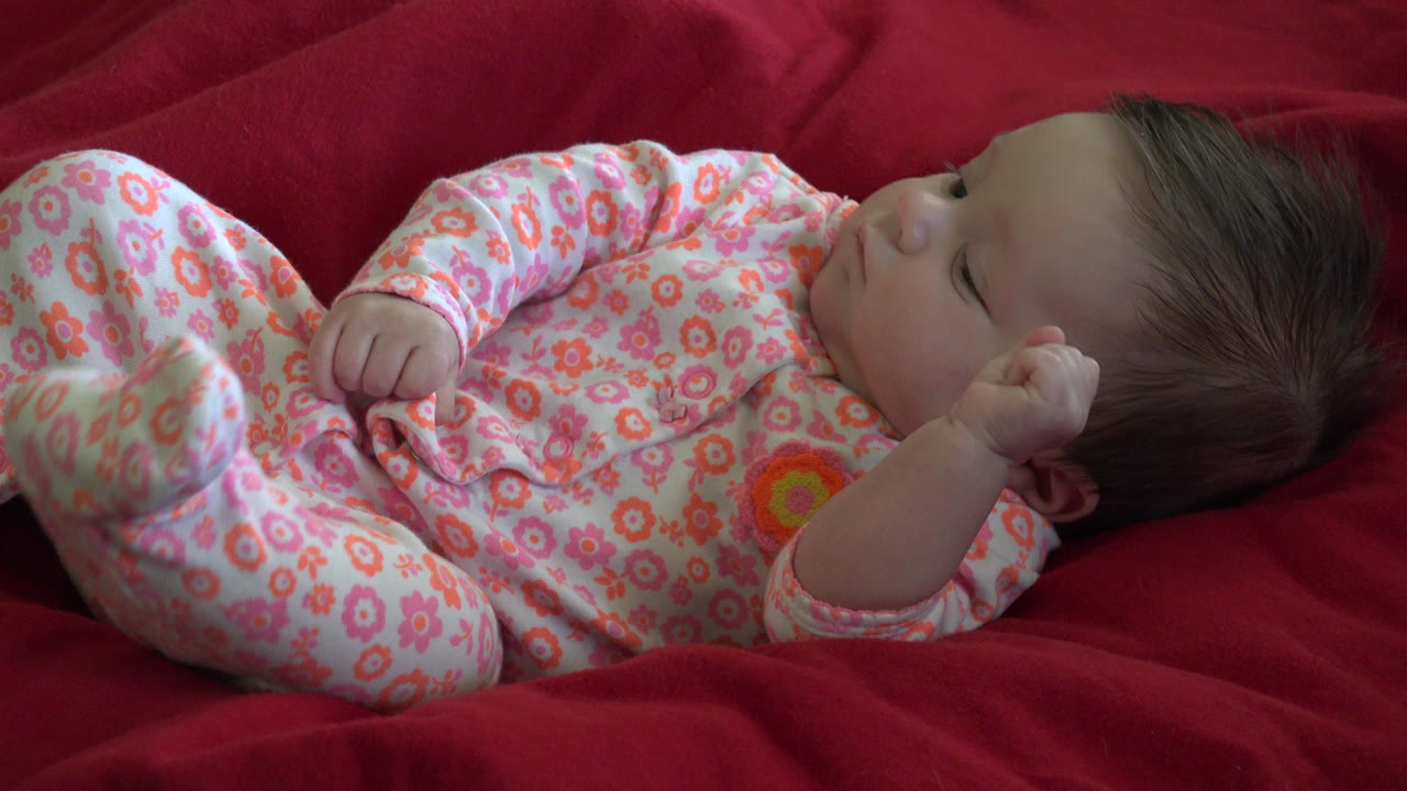 At the start of the Westminster, Colorado clip, a baby in a pink floral onesie lies on a red blanket with eyes mostly closed and one hand raised near the head.