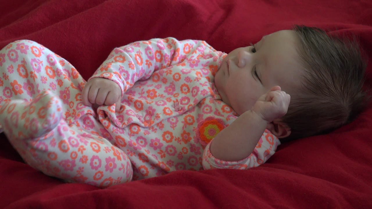 One second into the Westminster, Colorado footage, the baby remains reclined on the red blanket, wearing a pink floral outfit with a fist held near the chest.
