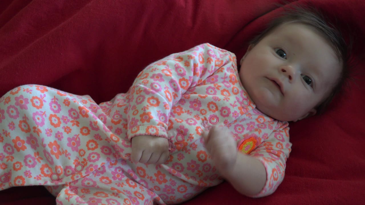 At 0:01, the baby remains on the red blanket in Westminster, Colorado, with one small hand slightly raised beside the floral sleeper.