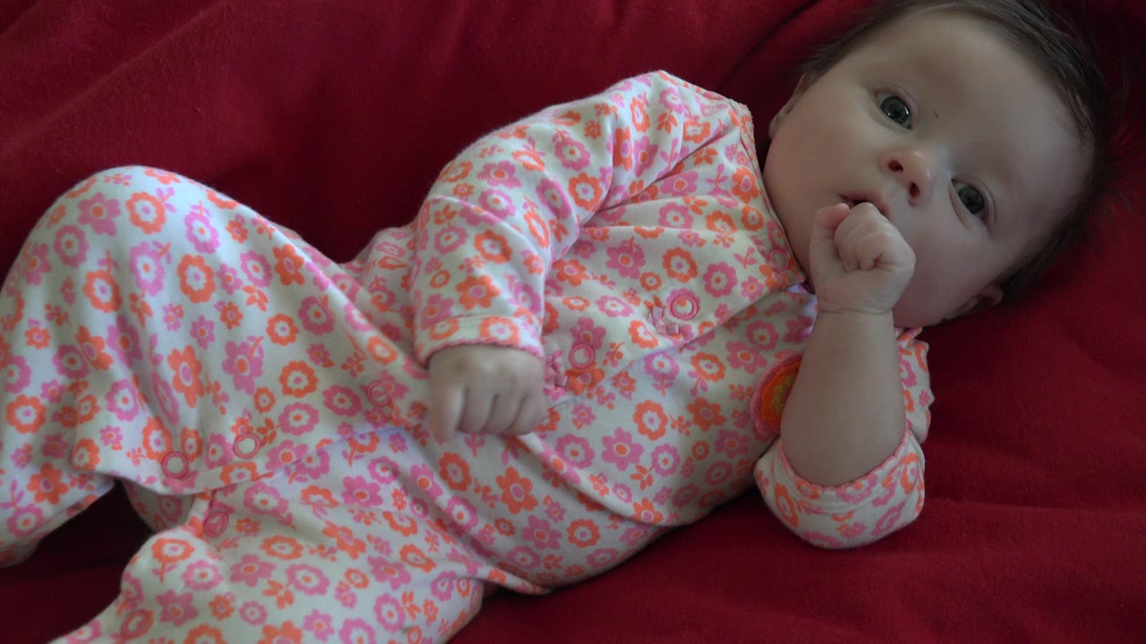 At 0:00, a baby in pink floral footed pajamas lies diagonally on a red couch in Westminster, Colorado, with one hand near the mouth.
