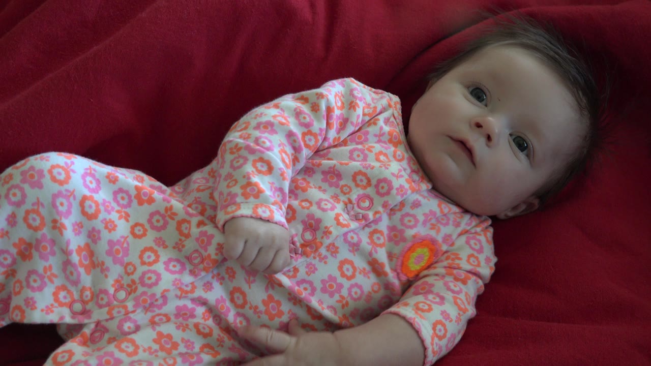 At 0:01, the baby remains on the red bedspread in Westminster, Colorado, with one hand curled near the floral sleeper and eyes open.