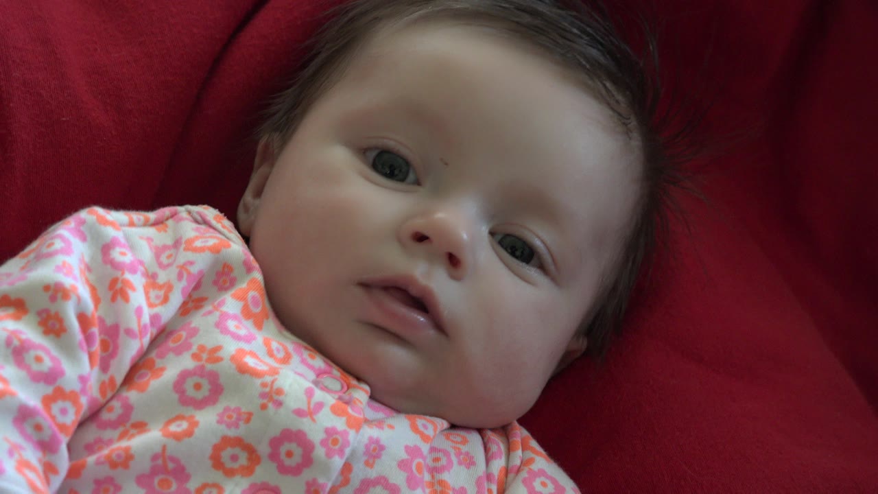 At 0:01, the awake infant remains on the red cushion with the face centered more fully and the floral outfit visible across the lower frame.