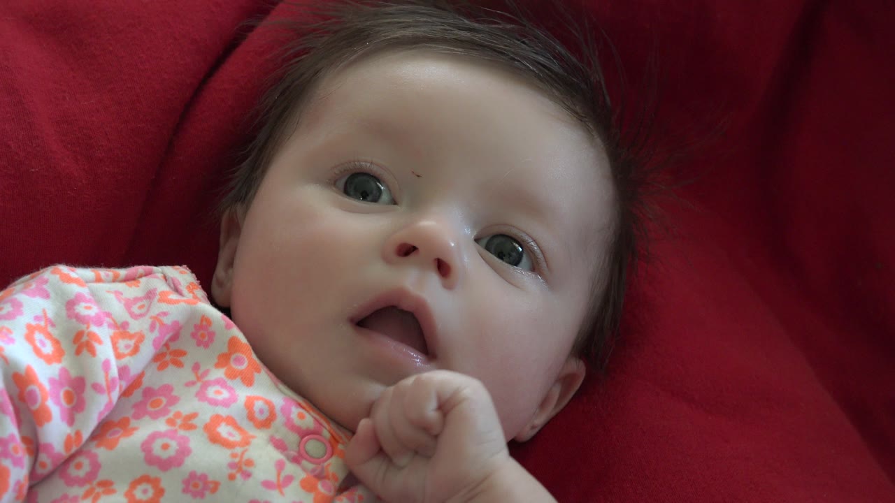 A close view of the baby in Westminster, Colorado shows the infant's hand raised near the mouth while lying on the red quilted blanket in a floral romper.