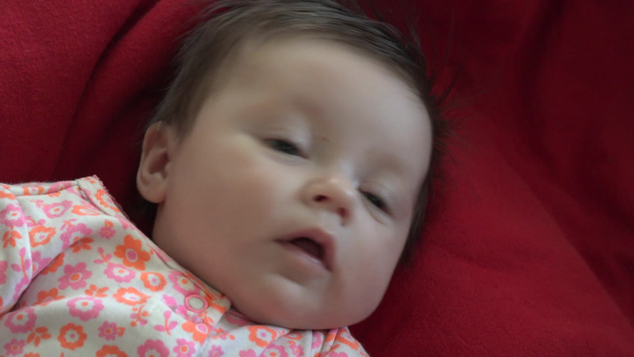 At 0:00, a baby in Westminster, Colorado lies close to camera on red fabric, wearing a pink floral outfit with eyes partly open and face angled toward the lens.