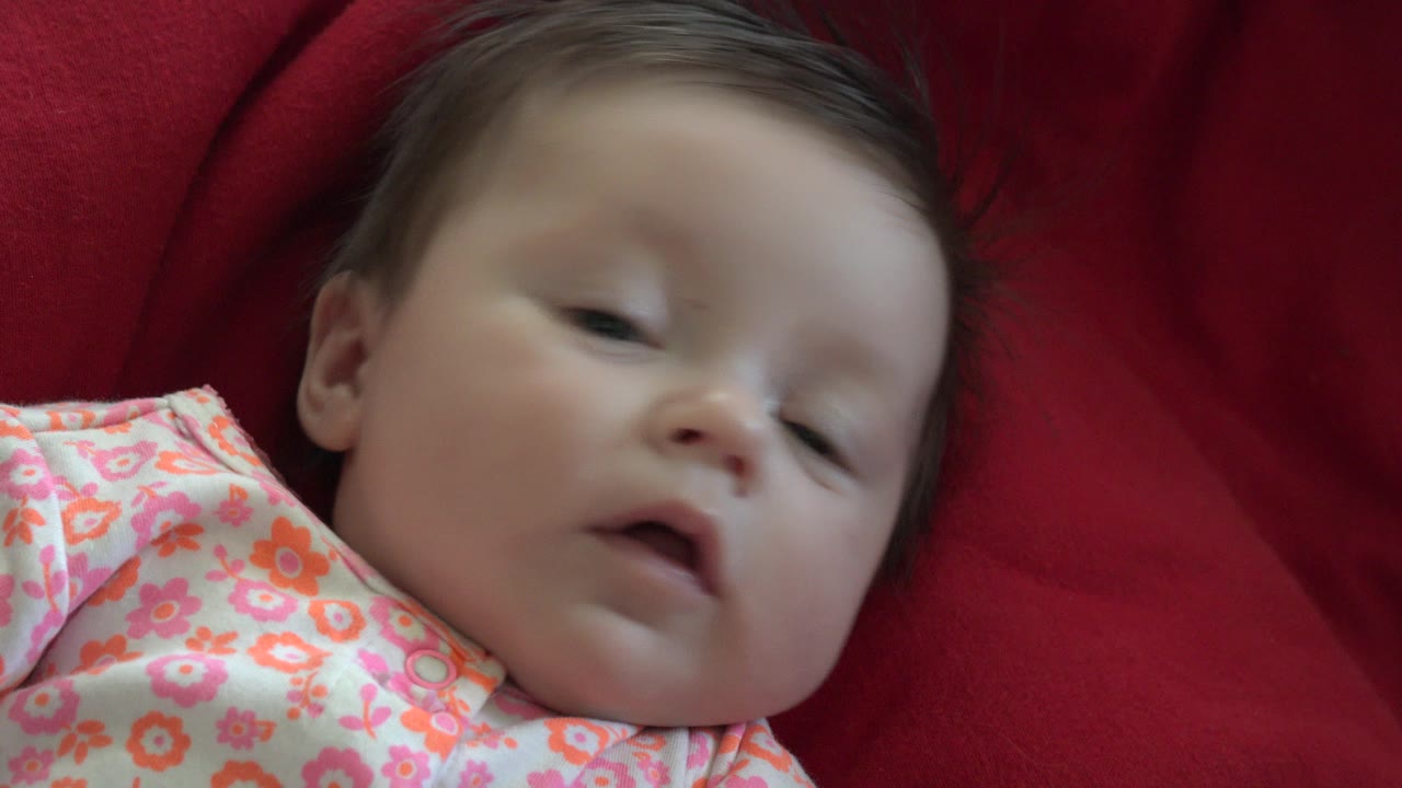 At 0:01, the baby remains on the red fabric in Westminster, Colorado, with eyes relaxed, mouth slightly open, and the floral clothing visible at the lower left.