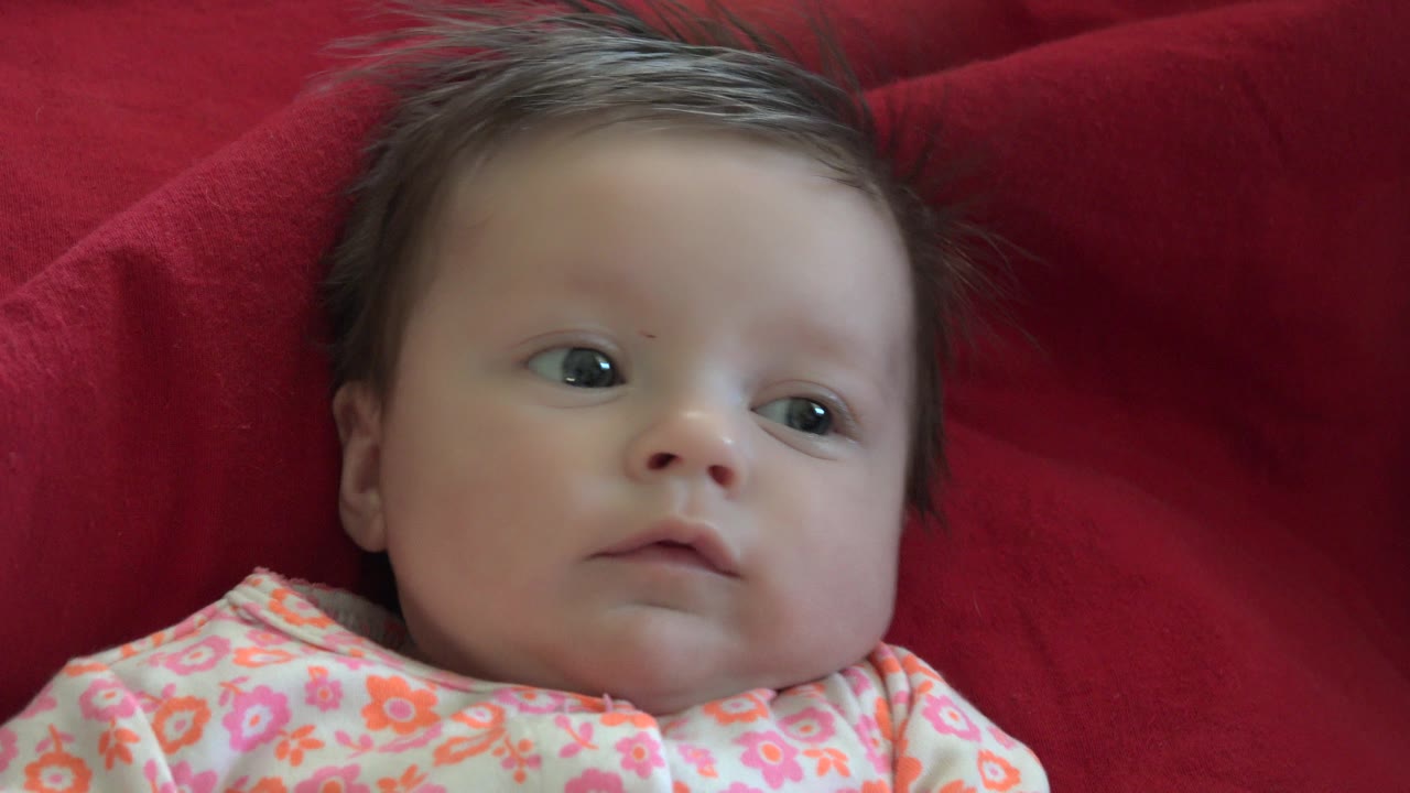 At 0:01, the baby remains on the red sofa or blanket, eyes turned off camera while the floral clothing fills the lower frame.