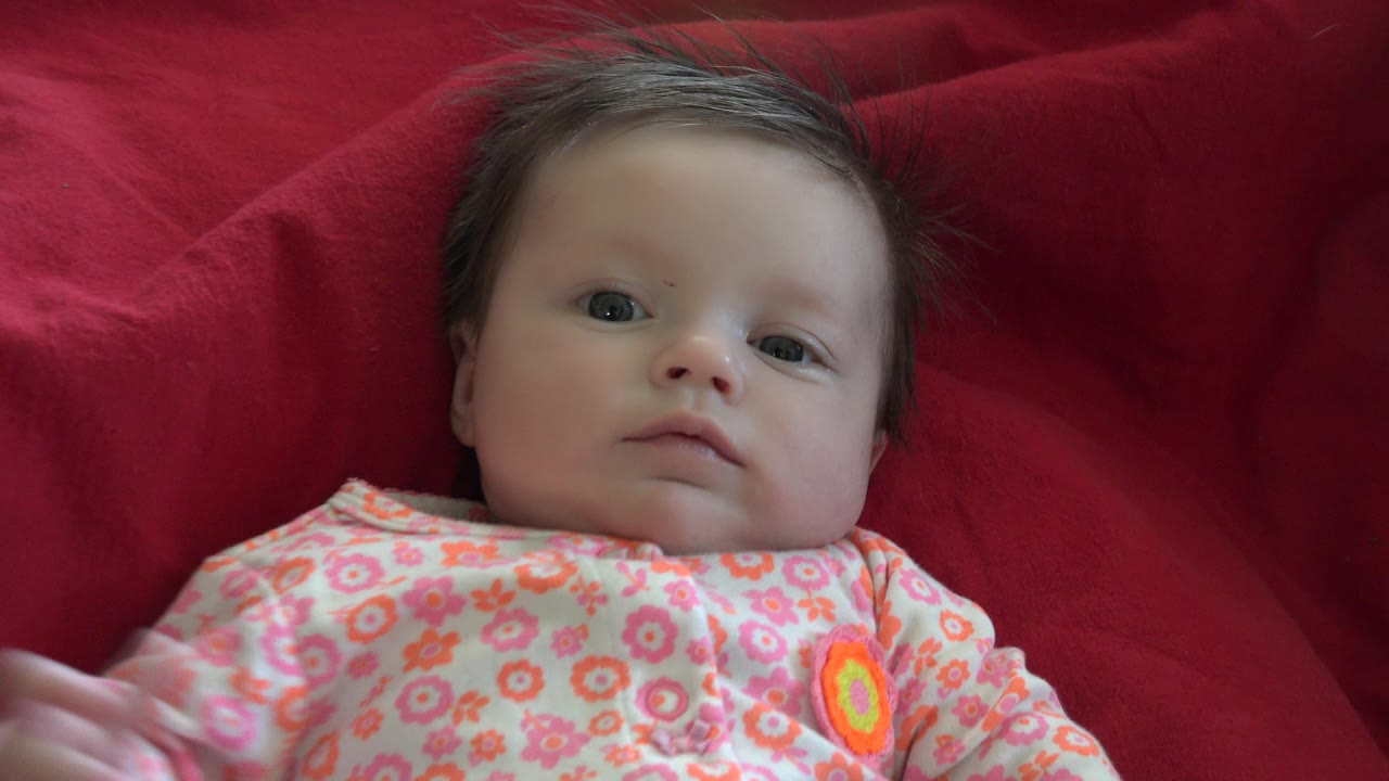 At 0:00, a baby in a pink floral outfit lies on a red blanket in Westminster, Colorado, looking toward the camera with one hand partly visible at the edge of frame.