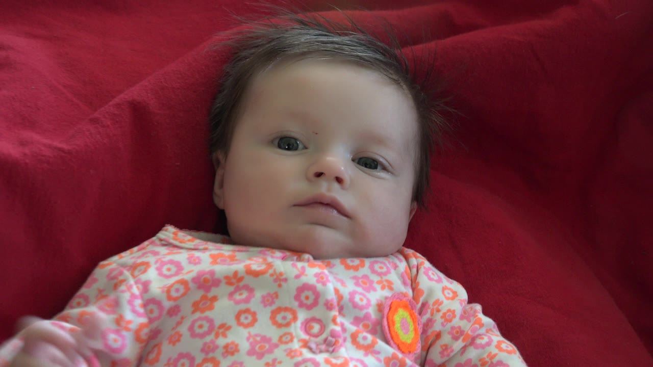 At 0:01, the baby remains centered on the red blanket in Westminster, Colorado, with eyes open, a neutral expression, and the floral outfit visible across the chest.