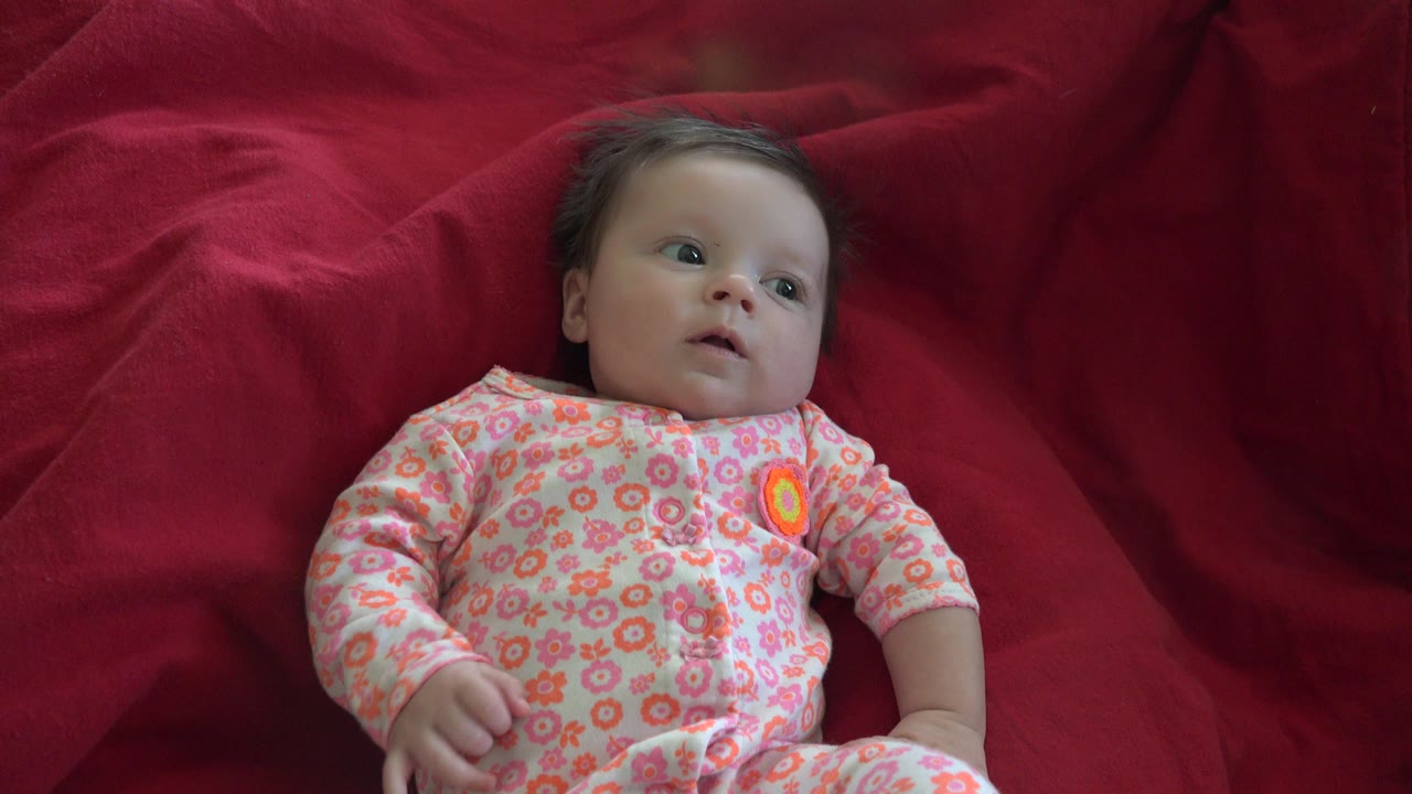 Baby in a pink floral onesie lying on a red blanket.