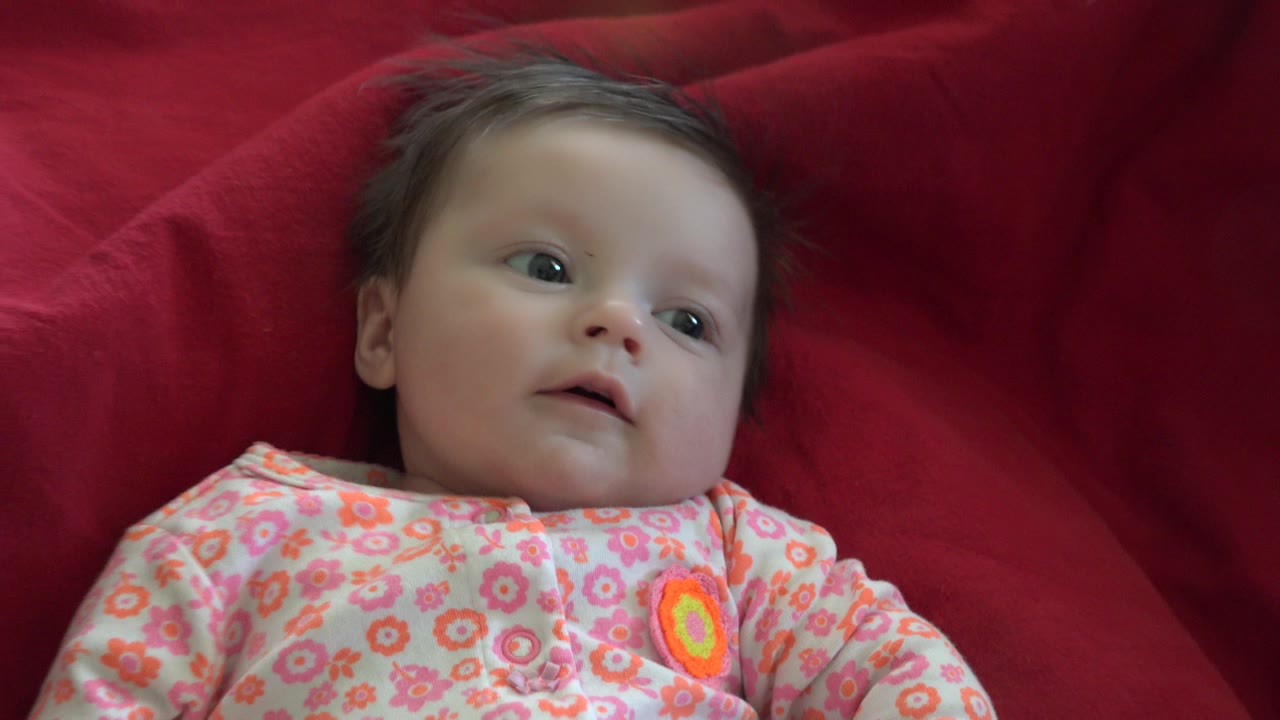 At 0:00, a baby in Westminster, Colorado lies on a red blanket in a pink floral outfit, looking upward and slightly to the side.