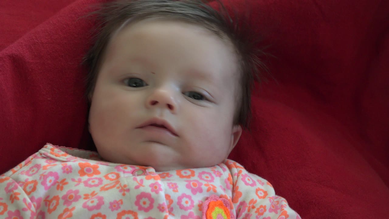 At 0:01, the baby remains centered on the red blanket in Westminster, Colorado, with eyes open and the patterned outfit filling the foreground.