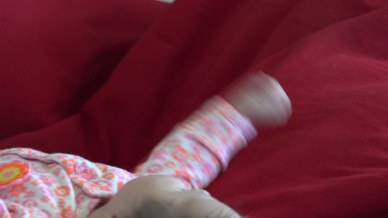 The baby remains close to the camera on the red cushion in Westminster, Colorado, with a raised leg blurred by movement and floral clothing filling the lower frame.